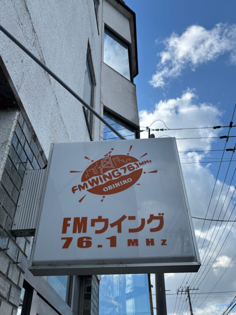 青空を背に、建物の壁に取りつけられたFMウイング 76.1MHzの看板